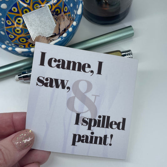 Hand holding a card with text 'I came, I saw, and I spilled paint!' near art supplies on a white background.